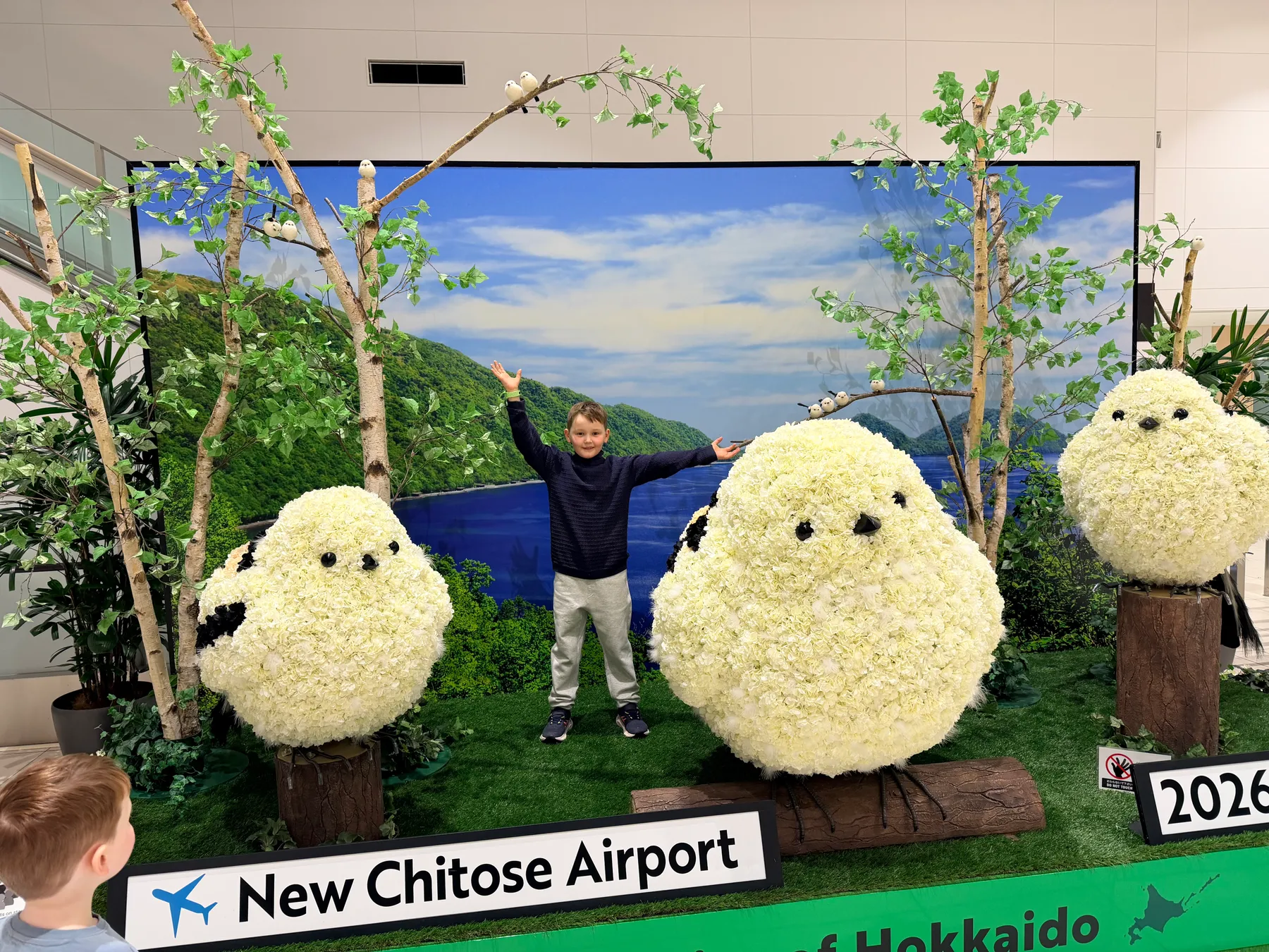 A child stretches their arms wide beside giant fluffy shima-enaga (long-tailed tit) flower sculptures at New Chitose Airport in Hokkaido.