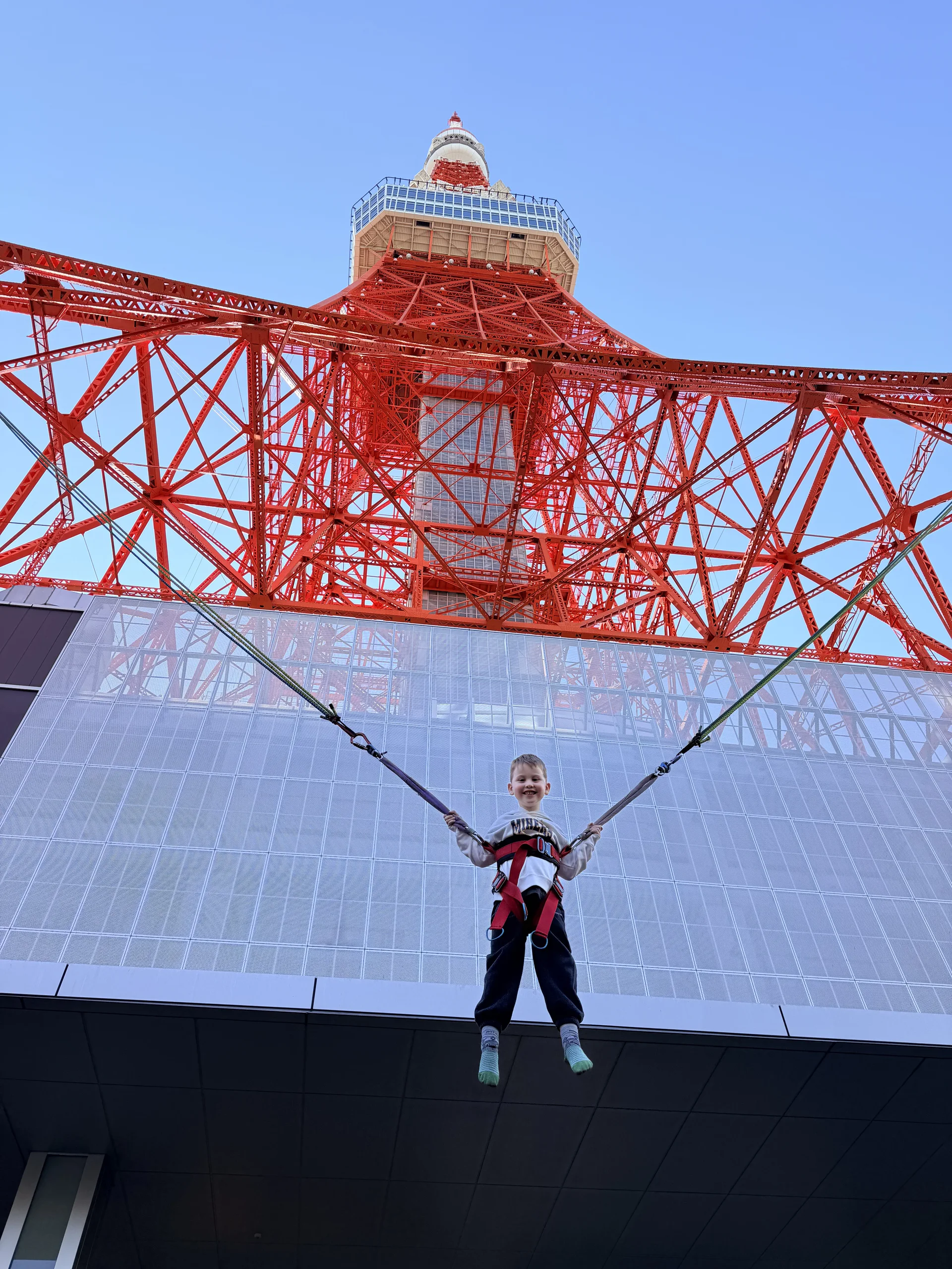 A child bouncing on a bungee-trampoline beneath the red steel framework of Tokyo Tower.