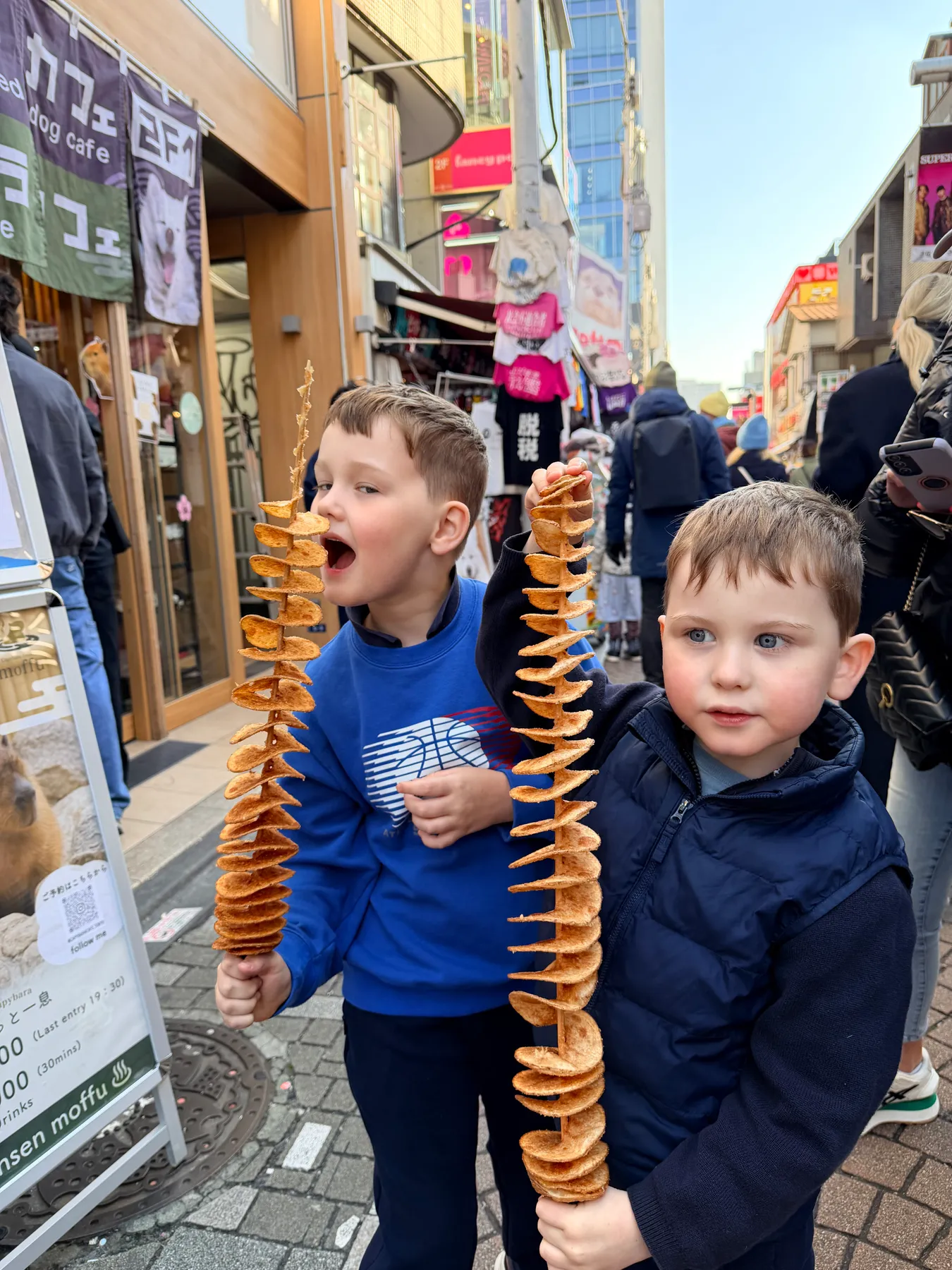 Two children hold spiral tornado-potato snacks on sticks on a busy Tokyo shopping street.